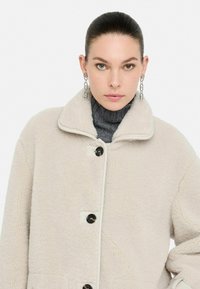 Una donna con i capelli all'indietro indossa orecchini a catena, un dolcevita grigio e un cappotto in sherpa color crema con grandi bottoni.