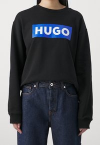 Black sweatshirt featuring a blue rectangular logo with the word "HUGO" in white. Fabric has a smooth texture, with long sleeves and a crew neckline.