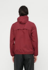 Chaqueta cortavientos burdeos con capucha y puños elásticos. Presenta una solapa en la parte posterior y un dobladillo ajustado para un aspecto entallado.
