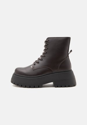Bottine marron foncé à lacets avec semelle noire épaisse et grosse et languette à l'arrière, vue de côté sur fond blanc.