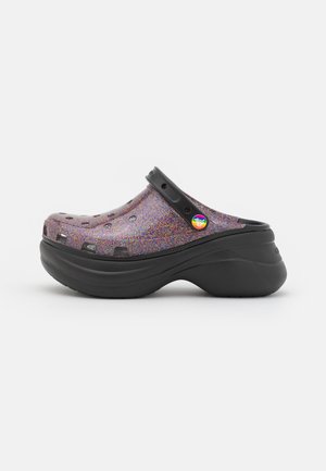 Sabot violet pailleté avec semelle plateforme noire épaisse et bride noire réglable avec un bouton arc-en-ciel fierté sur le côté.