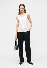 Mujer con top blanco sin mangas de escote en V, pantalones negros de pierna ancha, zapatos negros puntiagudos, sosteniendo un bolso negro y gris.