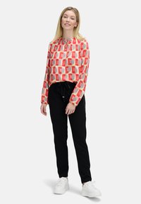 Langärmlige Bluse mit geometrischem rot-beigem Muster, kombiniert mit schwarzen, schmal geschnittenen Hosen und weißen Sneakers. Weicher, leichter Stoff.
