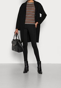 Svart lång kappa över en randig tröja i orange, vitt och brunt, tillsammans med svarta skinny jeans, ankelstövlar och en svart handväska.