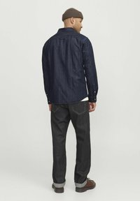 Camicia di denim scuro con maniche lunghe, cuciture azzurre, e colletto a punta, abbinata a jeans scuri e stivali marroni.