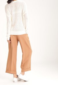 Nainen, joka on pukeutunut valkoiseen neulepaitaan, leveälahkeisiin camel-värisiin housuihin ja valkoisiin tennareihin, seisoo selkä kameraan päin.