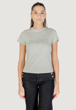 Giovane donna con capelli lunghi che indossa una maglietta Guess grigio chiaro a maniche corte e jeans scuri con bottoni laterali, in piedi contro uno sfondo bianco.