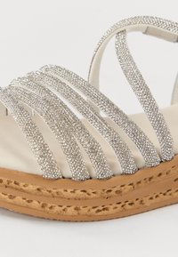 Silver strappy sandals featuring shimmering rhinestone embellishments, a cushioned beige insole, and a woven jute outsole.
