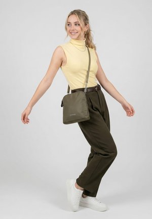 Junge Frau lächelt, trägt ein gelbes ärmelloses Oberteil, olivgrüne Hose, weiße Turnschuhe und trägt eine olivgrüne Umhängetasche.