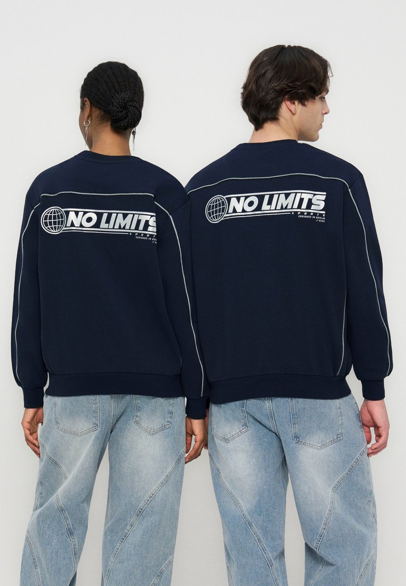 Δύο άτομα φοράνε ναυτικά φούτερ με άσπρο κείμενο «NO LIMITS» και λογότυπο γήινου σφαίρας στην πλάτη, συνοδευόμενα από ανοιχτόχρωμα μπλε τζιν.