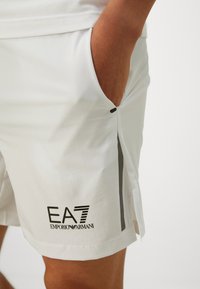 Osoba nosi bijele Emporio Armani EA7 kratke hlače s rukom u bočnom džepu, logo marke vidljiv blizu rubova.