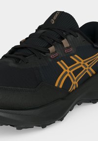 Chaussure de sport Asics noire et bleu foncé avec logo orange, lacets noirs et semelle en caoutchouc texturée visible depuis un angle latéral avant.
