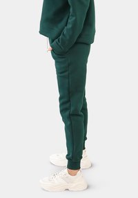 Grüne Baumwoll-Sweatpants mit elastischen Bündchen, Seitentaschen und einer lässigen Passform, kombiniert mit klobigen weißen Sneakers.