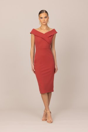 Γυναίκα που φοράει εφαρμοστή, κόκκινη φόρεμα μέχρι το γόνατο με ανοικτούς ώμους, μπεζ πέδιλα με λουράκι στον αστράγαλο, χρυσούς κρίκους σκουλαρίκια και χρυσό κολιέ, στέκεται μπροστά σε μπεζ φόντο.