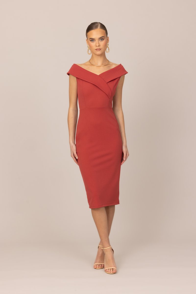 Γυναίκα που φοράει εφαρμοστή, κόκκινη φόρεμα μέχρι το γόνατο με ανοικτούς ώμους, μπεζ πέδιλα με λουράκι στον αστράγαλο, χρυσούς κρίκους σκουλαρίκια και χρυσό κολιέ, στέκεται μπροστά σε μπεζ φόντο.