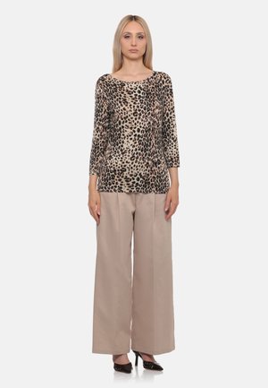 Donna in piedi che indossa un top a maniche lunghe con stampa leopardata, pantaloni beige a gamba larga e tacchi alti neri a punta su uno sfondo semplice.
