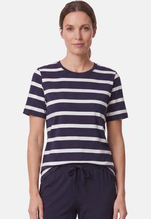 Frau trägt ein kurzärmeliges Shirt mit marineblauen und weißen horizontalen Streifen und marineblaue Hose mit Kordelzug, steht vor weißem Hintergrund.