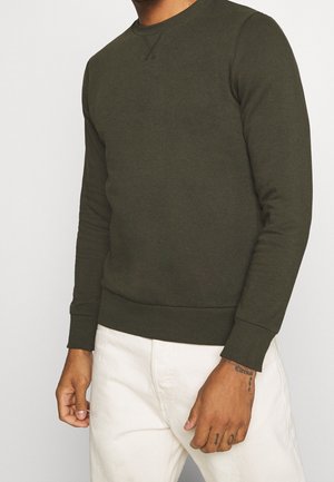 Mann trägt olivgrünen Langarmpullover mit Rundhalsausschnitt und weiße Hose, tätowierte Hände vor hellem Hintergrund sichtbar.