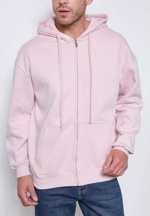 Homme portant un sweat à capuche zippé rose clair avec poches avant et cordons, assorti à un jean bleu, sur un fond uni.