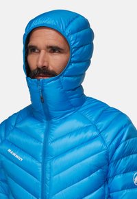 Veste isolante bleue avec capuche, présentant un design matelassé et une fermeture éclair avant. Le tissu a une texture lisse et brillante. Logo sur la manche.