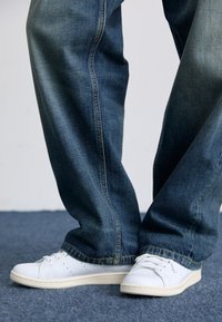 Zapatillas blancas con detalles perforados en los laterales, combinadas con vaqueros de denim azul oscuro con corte recto y costuras visibles.