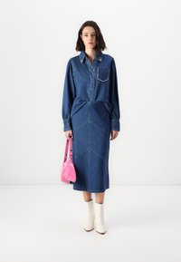 Jeanskjol med långa ärmar, fickor och dekorativ sömnad; matchad med en rosa handväska och vita ankelskor, framför en enfärgad bakgrund.