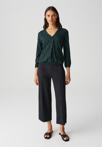 Top nero a maniche lunghe con motivo astratto verde, scollatura a V e orlo arricciato, abbinato a pantaloni neri a gamba larga e sandali piatti.