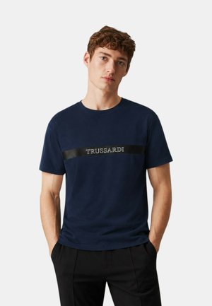 Giovane uomo con capelli ricci che indossa una T-shirt blu navy con una striscia nera e la scritta bianca "TRUSSARDI", mani in tasca, sfondo chiaro.