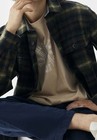 Camisa de franela a cuadros en verde oscuro y azul marino, superpuesta a una camiseta gráfica beige con texto circular. Combinada con pantalones azul marino.