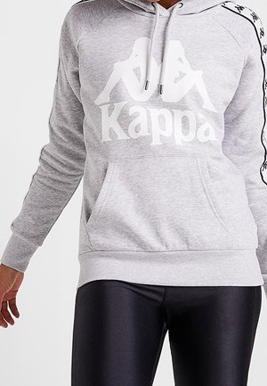 Grå Kappa hættetrøje med hvidt logo af to siddende figurer ryg mod ryg, båret med sorte skinnende leggings på en person mod en hvid baggrund.
