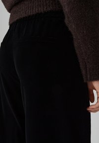 Zwarte fluwelen broek met een elastische tailleband, voorzien van twee achterzakken, afgebeeld naast een gestructureerde bruine trui. Close-up van de zijkant.