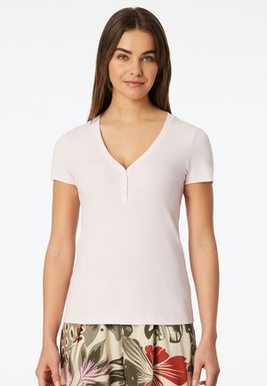 Woman with long brown hair wearing a light pink short-sleeve V-neck top and floral patterned pants standing against a white background.