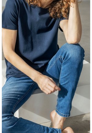 Osoba o kręconych włosach, ubrana w granatowy t-shirt i niebieskie dżinsy, siedząca boso z jedną uniesioną kolanem i opartą na nim ręką.