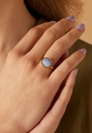 Gold ring featuring a smooth, oval blue stone. The ring has a thin band and a beveled edge around the stone. Light skin tone and pale purple nails.