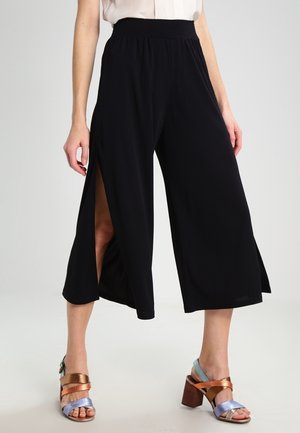 Vrouw die zwarte, wijde, cropped broek met zijopeningen draagt en multikleurige sandalen met hoge hakken, staande tegen een effen achtergrond.