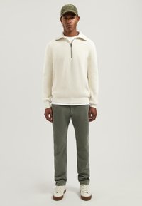 Crème ribgebreide trui met ritssluiting, olijfgroene broek en witte sneakers. De hoed is donkergroen, wat de casual outfit compleet maakt.