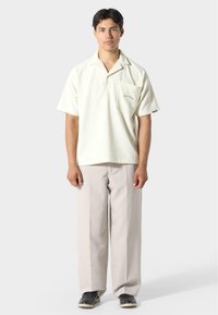 Polo tričko s krátkým rukávem v světlé béžové barvě s texturovanou tkaninou a jednou náprsní kapsou, spárované s širokými kalhotami světle šedé barvy a černými botami.