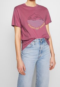 Mörkröd bomull t-shirt med en svart späckhuggargrafik och gul text "Skydda våra hav," i kombination med ljusblå högmidjade jeans.