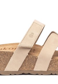 Sandale en daim beige avec deux larges brides, semelle intermédiaire en liège et semelle extérieure en caoutchouc texturé. Présente un logo en relief sur la bride et un motif naturel en liège.
