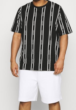Man die een zwart shirt met korte mouwen draagt met verticale witte strepen en witte korte broek tegen een effen lichte achtergrond.