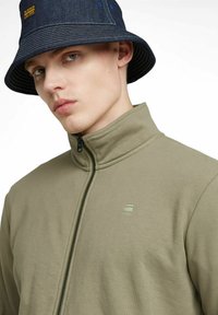 Giacca a zip verde oliva con colletto alto, texture morbida e piccolo logo sul petto sinistro; abbinata a un cappello a secchiello in denim.
