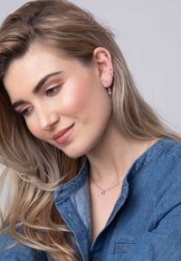 Zilveren ketting met een kleine paarse edelsteen, gecombineerd met meerdere oorbelgaatjes en gestyled met lang, golvend blond haar. Spijkerblouse.