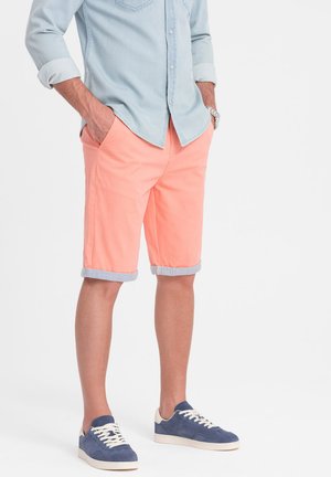 Shorts de couleur pêche avec des ourlets repliés, fabriqués en tissu léger. Associés à une chemise en denim bleu clair et des baskets navy.