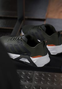 Paire de baskets athlétiques vert foncé avec des détails noirs, blancs et orange reposant sur une surface sombre avec un arrière-plan flou.