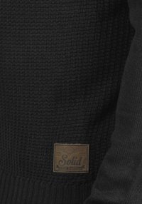 Maglione nero lavorato a maglia con tessuto testurizzato. Presenta una toppa in pelle marrone con il logo "Solid" sul lato sinistro in basso.