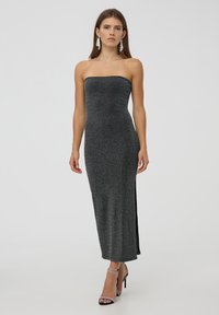 Abito aderente senza spalline di colore nero con riflessi metallici. Presenta una fessura laterale e un design lungo fino al pavimento. Abbinato a sandali con cinturino e tacco alto.