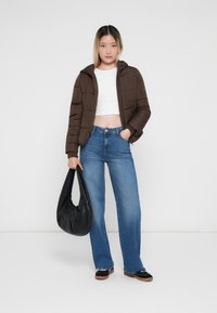 Veste matelassée marron avec capuche, top blanc court, jeans bleu à jambes larges et sac à main noir. Tissu texturé et couleurs contrastées.