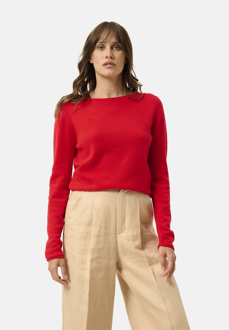 Donna con capelli castani ondulati che indossa un maglione rosso a maniche lunghe e pantaloni beige a vita alta e larghi, in piedi davanti a uno sfondo bianco.