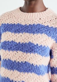 Gros plan d'une personne portant un pull en maille texturée à larges rayures alternées en rose clair et bleu pervenche.