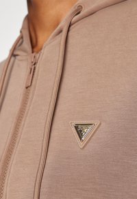 Felpa zip-up marrone chiaro realizzata in tessuto morbido, con un accento logo triangolare in oro e beige vicino alla cerniera.